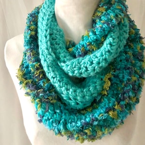 Op de afbeelding: Een tealblauwe gecrochete oneindige sjaal met een pluche, veelkleurige garen accent. De sjaal is gemaakt met een dik garen en heeft een eenvoudig, elegant ontwerp.