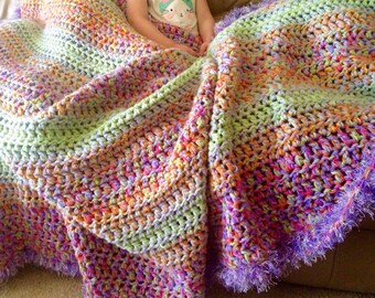 May include: A multi-colored crocheted blanket with a purple fringe. The blanket is made with a variety of colors, including green, orange, pink, and purple.