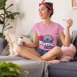 Może przedstawiać: Różowa koszulka z napisem "My Little Panic Attacks" i tęczą. Osoba nosząca koszulkę siedzi na szarej sofie. Ma również na sobie dżinsowe szorty i czerwoną opaskę na głowę.
