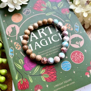 May include: A green book with the title "Earth Magick" and illustrations of flowers, mushrooms, and a cat. A wooden and stone beaded bracelet with a copper charm is on top of the book.