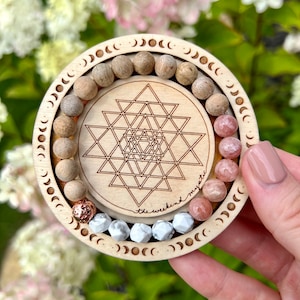 May include: A wooden bracelet with a geometric design, surrounded by a circle of moon phases. The bracelet is made of natural wood beads and features a pink and white stone design. The text "the weekend mystic" is engraved on the wooden circle.