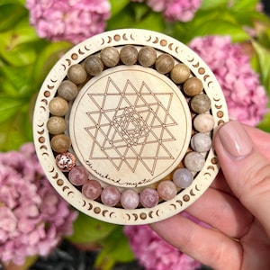 Può includere: Un cerchio di legno con un design di fasi lunari attorno al bordo. Il cerchio ha un design geometrico al centro e un braccialetto di perline rosa e marroni all'interno. Il braccialetto ha il testo "the weekend mystic" su di esso.