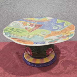 May include: A decorative ceramic cake stand with a colorful, abstract design on the plate. The plate has a scalloped edge and rests on a pedestal base with floral and geometric patterns. The base has a dark green center with red flowers.