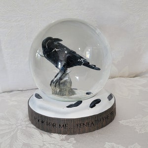May include: A snow globe featuring a black crow perched on a tree stump inside a clear glass sphere. The base is brown with the text "LOOK FOR ME... BENEATH THE TREE". Black feather accents surround the base.
