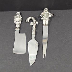 May include: Three silver-toned cheese knives with decorative handles. One knife features a nutcracker, another a candy cane, and the third a Santa Claus figure. The blades are stainless steel. The knives are arranged on a dark surface.