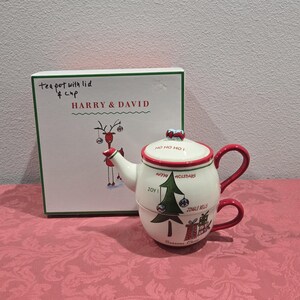 May include: A white ceramic Christmas teapot with a red handle and trim, stacked on a matching cup. The teapot features a Christmas tree design and the words "Happy Holidays" and "Jingle Bells". The box reads "HARRY & DAVID".