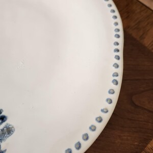 May include: A white ceramic plate with a blue dotted border. The plate has a slightly irregular edge and a simple, elegant design. The plate is likely for serving food.