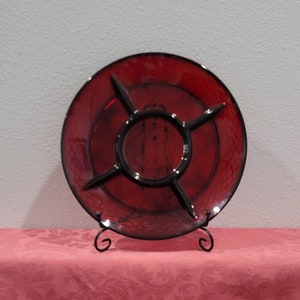 May include: A red glass serving dish with four sections. The dish has a raised, textured design and is on a black metal stand.