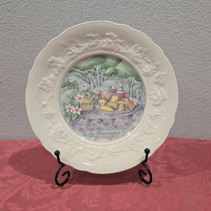 May include: White decorative plate with a raised grape vine design around the rim. The center features a colorful illustration of a table set with cheese, wine, and flowers. The plate is displayed on a black metal stand.