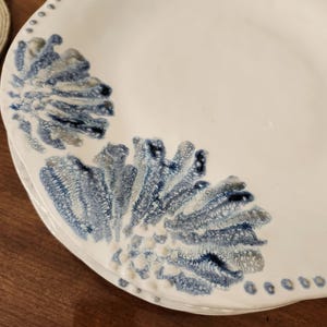 May include: White ceramic plates with a blue and gray floral design. The plates have a scalloped edge with small dotted details. The design is reminiscent of coral or sea plants.
