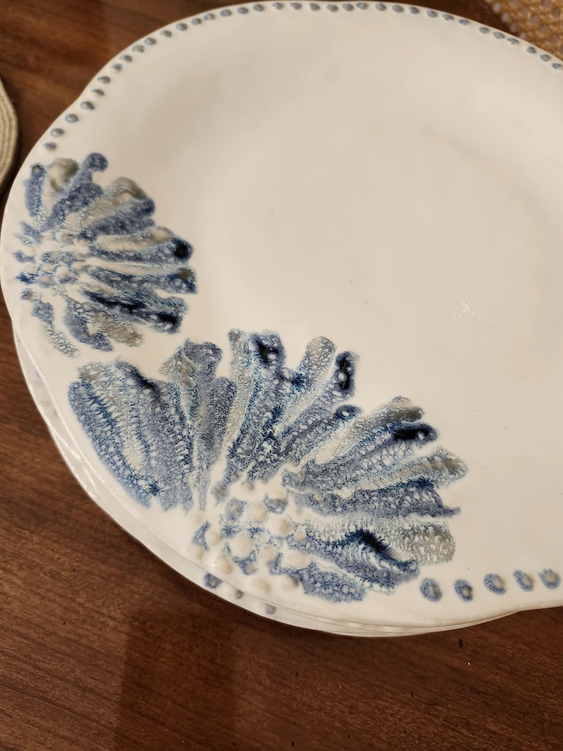 May include: White ceramic plates with a blue and gray floral design. The plates have a scalloped edge with small dotted details. The design is reminiscent of coral or sea plants.