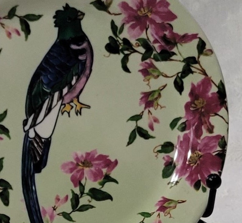 May include: A green and blue bird with a long tail sits on a pale green plate with pink flowers. The plate has a floral design with pink flowers and green leaves.