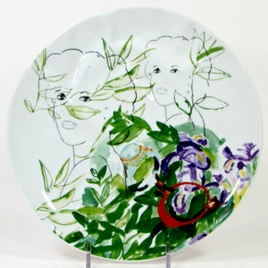 Puede incluir: Plato de cer&aacute;mica blanco con un dise&ntilde;o pintado a mano que presenta dos rostros de mujeres rodeados de hojas verdes y flores moradas.