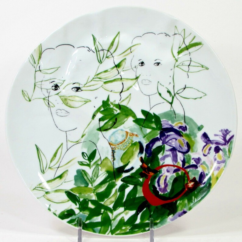 Puede incluir: Plato de cer&aacute;mica blanco con un dise&ntilde;o pintado a mano que presenta dos rostros de mujeres rodeados de hojas verdes y flores moradas.