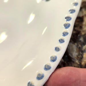 May include: Close-up of a white ceramic plate with a blue floral design and a beaded edge. The edge features small, evenly spaced blue dots. The plate is partially visible, showcasing its curved shape and glossy finish.