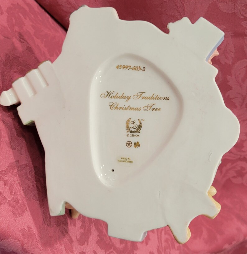 Op de afbeelding: Witte porseleinen kerstboomversiering met gouden opschrift "Holiday Traditions Christmas Tree" en het Lenox-logo. De versiering heeft productnummer 45997-605-2.