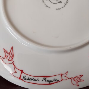 Puede incluir: Plato de cer&aacute;mica blanco con una cinta roja y dos p&aacute;jaros rojos. La cinta tiene el texto "Rebekah Mayello" escrito en tinta negra.