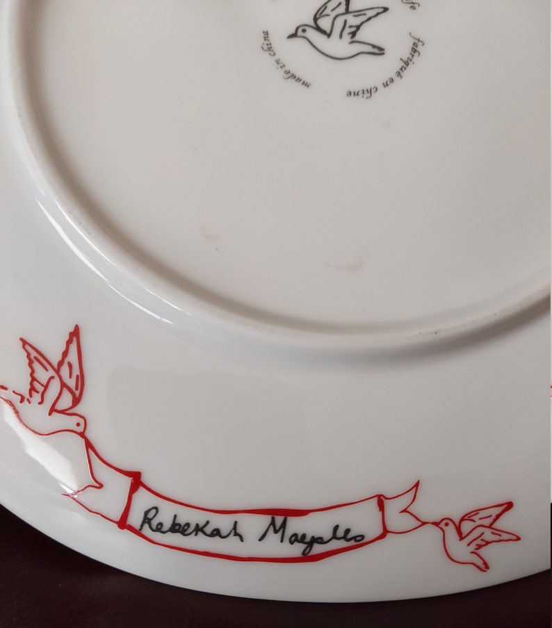 Puede incluir: Plato de cer&aacute;mica blanco con una cinta roja y dos p&aacute;jaros rojos. La cinta tiene el texto "Rebekah Mayello" escrito en tinta negra.