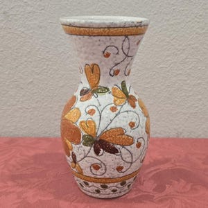 May include: A white ceramic vase with a textured surface, adorned with orange, brown, and green floral patterns. The vase has a flared top and a decorative band around the rim and base. The vase is approximately 7 inches tall.