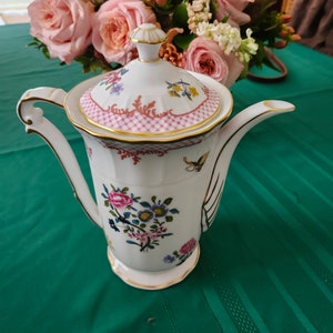 May include: A white porcelain coffee pot with a floral design and gold accents. The lid has a pink and gold lattice pattern. The pot is sitting on a green tablecloth.