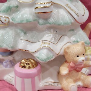Op de afbeelding: Een witte porseleinen kerstboomfiguur met gouden accenten. De boom is versierd met een roze cadeaudoos, een gouden lint en een teddybeer. De namen "Amanda" en "Josie" staan op de boom.