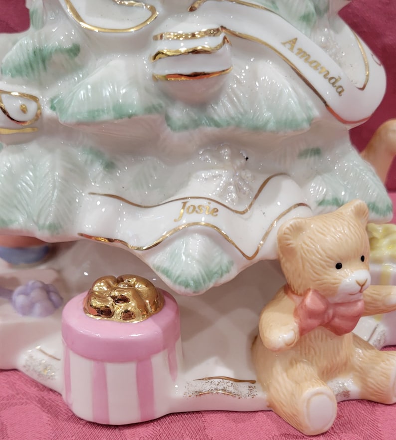 Op de afbeelding: Een witte porseleinen kerstboomfiguur met gouden accenten. De boom is versierd met een roze cadeaudoos, een gouden lint en een teddybeer. De namen "Amanda" en "Josie" staan op de boom.