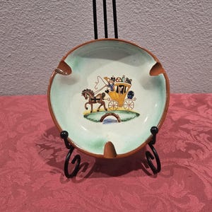 May include: A round ceramic ashtray with a light green interior and brown rim. The center features a colorful illustration of a horse-drawn carriage on a bridge. The ashtray has three curved indentations for resting cigarettes.