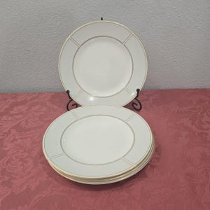 May include: A set of white ceramic plates with a light gray border and gold trim. The plates are stacked and displayed on a red tablecloth. One plate is propped up on a black metal stand.
