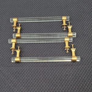 May include: A set of four cabinet pulls featuring clear, cylindrical acrylic bars connected to metallic gold-toned square mounting brackets. Each pull includes two matching gold-toned screws positioned at the base of the mounting hardware.