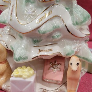 Op de afbeelding: Een witte porseleinen kerstboomversiering met groene bladeren en gouden versiering. De versiering heeft drie namen: "Candace", "Jessica" en "Miranda". De versiering bevat ook een klein roze boek, een gele geschenkdoos en een klein bruin paardenbeeldje.
