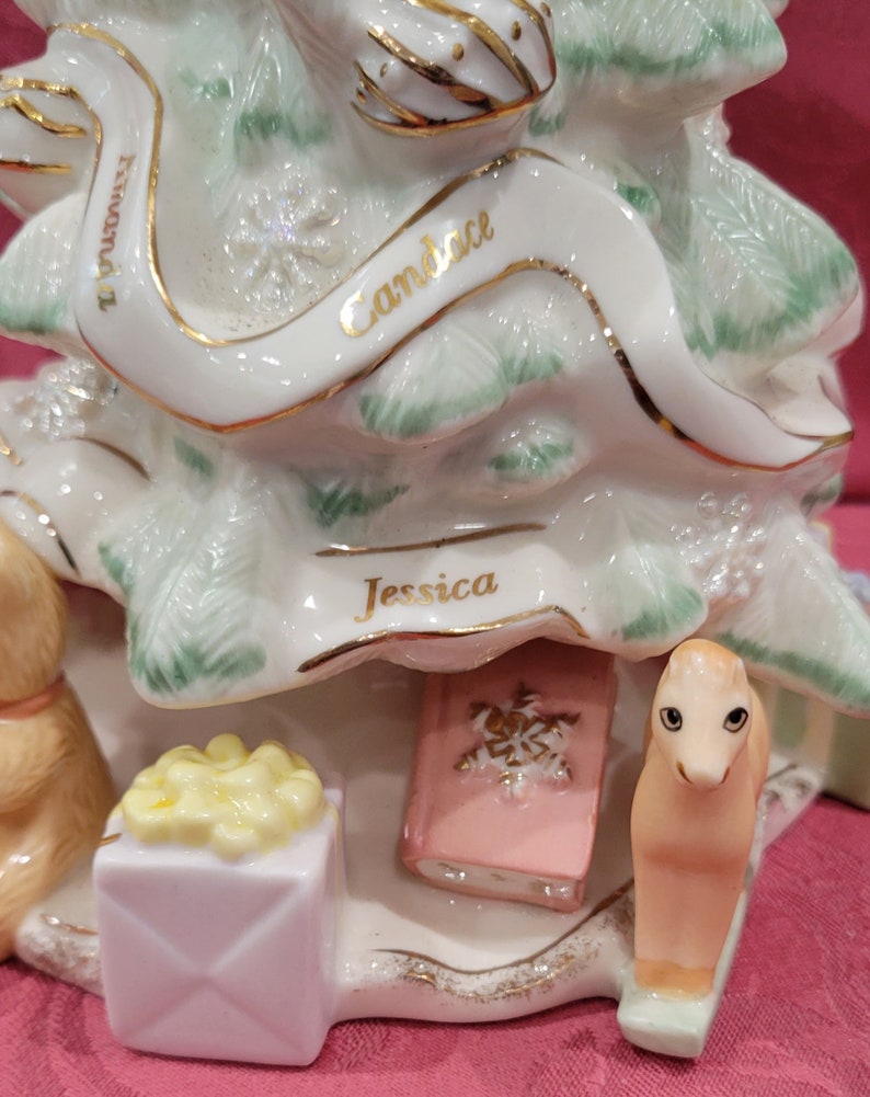 Op de afbeelding: Een witte porseleinen kerstboomversiering met groene bladeren en gouden versiering. De versiering heeft drie namen: "Candace", "Jessica" en "Miranda". De versiering bevat ook een klein roze boek, een gele geschenkdoos en een klein bruin paardenbeeldje.