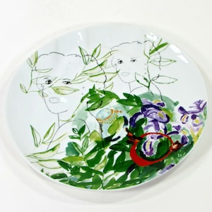 Puede incluir: Un plato de cer&aacute;mica blanco con un dise&ntilde;o floral verde, morado y rojo. Dos dibujos en blanco y negro de rostros de mujeres est&aacute;n parcialmente ocultos por el dise&ntilde;o floral.