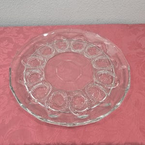 May include: Clear glass egg serving platter with a circular design. The platter features a central octagonal area surrounded by a ring of egg-shaped indentations. The outer rim has a scalloped edge.