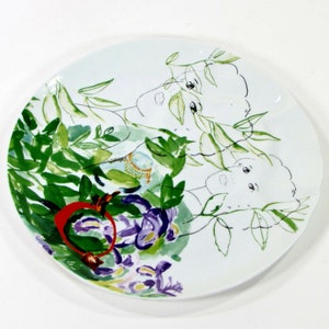 Puede incluir: Plato de cer&aacute;mica blanco con un dise&ntilde;o pintado a mano de dos caras rodeadas de hojas verdes y flores moradas. Las caras est&aacute;n delineadas en negro y tienen un efecto acuarela.
