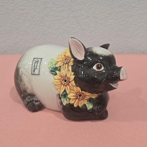 May include: A ceramic pig figurine with a black head and legs, and a white body. The pig is adorned with a yellow sunflower garland around its neck. The words "Hand Painted" are on the side. The figurine is on a pink surface.