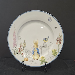 May include: White ceramic plate with a blue rim, featuring a charming illustration of Peter Rabbit in a blue jacket, surrounded by flowers, a bird, and a watering can. A butterfly and bee are also depicted. The plate is displayed on a black metal stand.