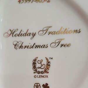 Op de afbeelding: Een witte Lenox kerstboomversiering met gouden opschrift "Holiday Traditions Christmas Tree" en een gouden Lenox-logo. De versiering heeft ook een gouden nummer 45997-605-2.