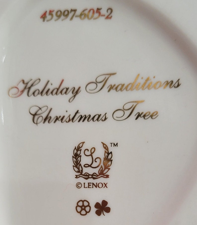 Op de afbeelding: Een witte Lenox kerstboomversiering met gouden opschrift "Holiday Traditions Christmas Tree" en een gouden Lenox-logo. De versiering heeft ook een gouden nummer 45997-605-2.
