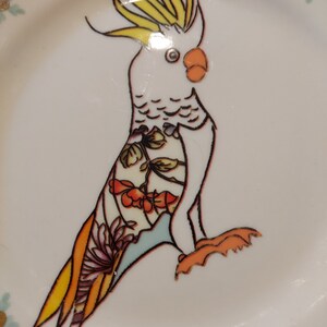 May include: A white ceramic plate with a hand-painted design of a cockatoo with yellow and orange accents. The cockatoo is perched on a branch with green and blue leaves. The plate has a white background with gold accents.