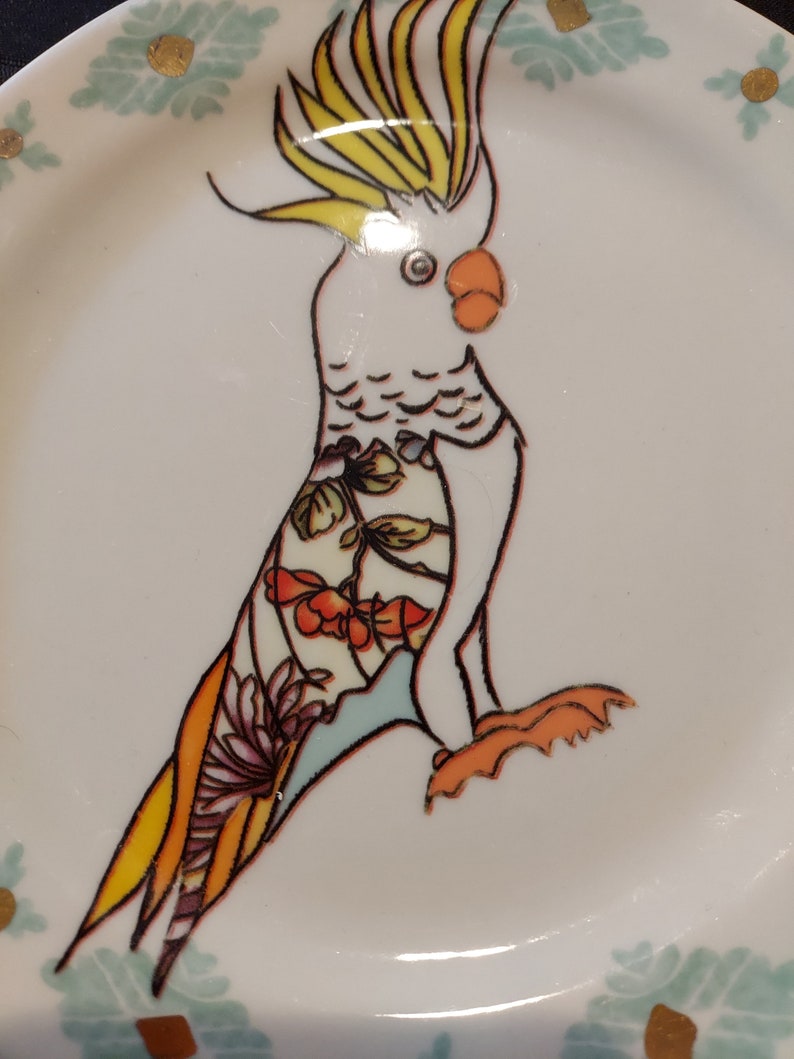 May include: A white ceramic plate with a hand-painted design of a cockatoo with yellow and orange accents. The cockatoo is perched on a branch with green and blue leaves. The plate has a white background with gold accents.