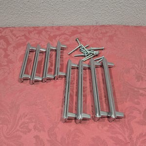 May include: A set of ten silver-toned drawer pulls with a sleek, modern design. The pulls are arranged in two rows of five, with each pull featuring a slightly curved, rectangular shape.