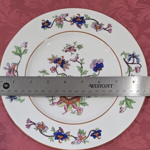 May include: A white ceramic plate with a gold rim and a floral design. The flowers are pink, blue, and green. The plate is 9 inches in diameter.