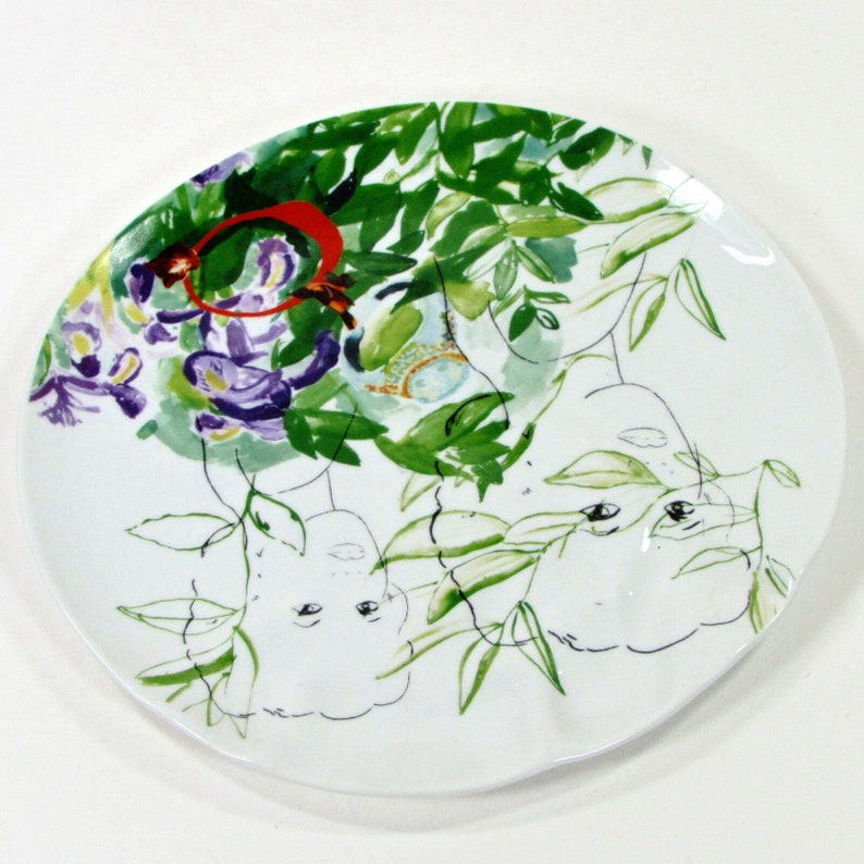 Puede incluir: Un plato de cer&aacute;mica blanco con un dise&ntilde;o pintado a mano de hojas verdes, flores moradas y dos caras. Las caras est&aacute;n parcialmente ocultas por las hojas y las flores.