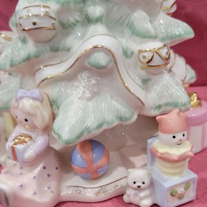 Op de afbeelding: Een witte porseleinen kerstboomfiguur met een groene glazuur. De boom is versierd met gouden versieringen en ornamenten. Er zijn ook figuurtjes van een meisje dat een cadeau vasthoudt, een sneeuwpop in een doos en een teddybeer.