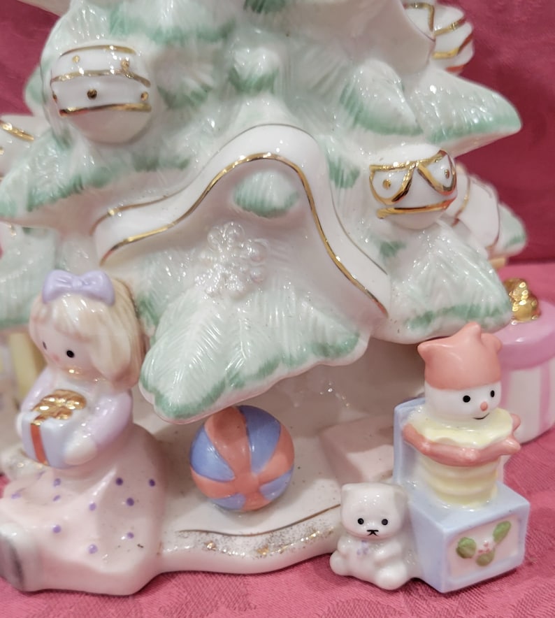 Op de afbeelding: Een witte porseleinen kerstboomfiguur met een groene glazuur. De boom is versierd met gouden versieringen en ornamenten. Er zijn ook figuurtjes van een meisje dat een cadeau vasthoudt, een sneeuwpop in een doos en een teddybeer.