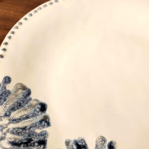 May include: A white ceramic plate with a decorative blue and gray coral-like design. The plate has a slightly scalloped edge with small, evenly spaced blue dots around the rim. The design is concentrated in the lower left corner.