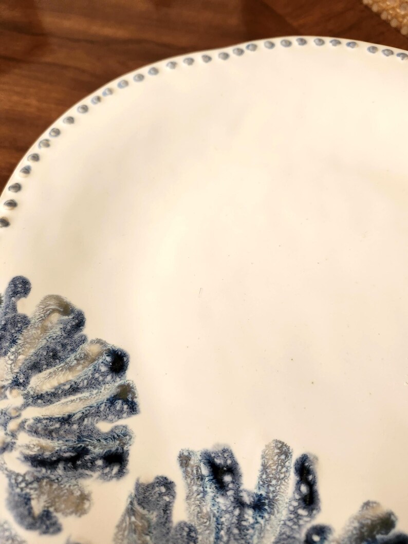 May include: A white ceramic plate with a decorative blue and gray coral-like design. The plate has a slightly scalloped edge with small, evenly spaced blue dots around the rim. The design is concentrated in the lower left corner.