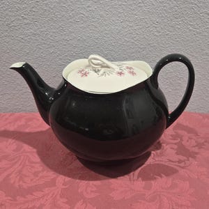 May include: A black ceramic teapot with a curved handle and spout. The lid is white with a floral pattern in pink and gray. The teapot is sitting on a pink tablecloth.