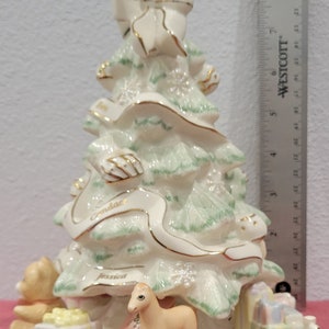 Op de afbeelding: Een witte porseleinen kerstboomfiguur met groene en gouden accenten. De boom is versierd met een witte strik, een gouden lint en de namen "Candace" en "Jessica". De basis van de boom heeft een teddybeer, een schommelpaard en ingepakte cadeaus.