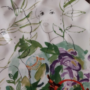 Puede incluir: Un plato de cer&aacute;mica blanco con un dise&ntilde;o floral estilizado que presenta un rostro de mujer, hojas verdes y flores moradas. El plato tiene un borde ondulado.